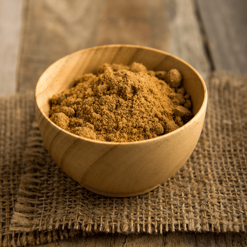 A smooth wooden bowl filled with fine, unrefined premium palmyra palm sugar. The bowl sits on a rustic woven burlap cloth over a wooden table.