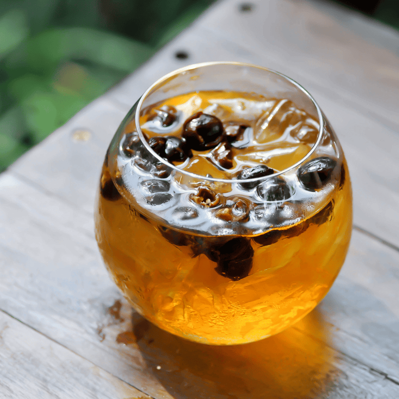Refreshing iced Fine Robusta cascara tea from Lamae Coffee. A cold, golden infusion over ice, garnished with rehydrated coffee cherry husks resting on a rustic wooden surface.