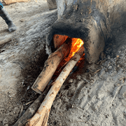 A traditional outdoor earthen stove with a bright, hot fire burning inside. Long wooden logs are being fed into the kiln's opening to boil and process unrefined palmyra palm sugar.